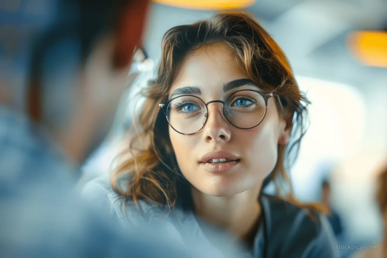 A young woman in glasses speaks during a team meeting in a bright office.