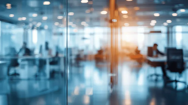 Modern office with glass walls and blurred workers at desks, showcasing a bright and productive workspace.