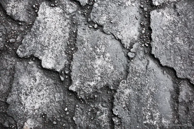 Close-up view of cracked grey asphalt pavement with loose gravel scattered in uneven damaged patches.