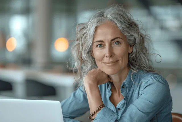 Mature woman with grey hair smiles while working on a laptop at a desk in a modern office.