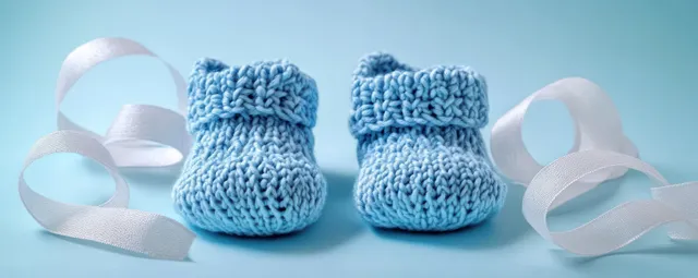 Pastel blue knitted baby shoes with white ribbon loops on soft blue studio background