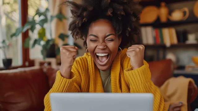 Joyful Black woman celebrating career success at home, cheering with laptop