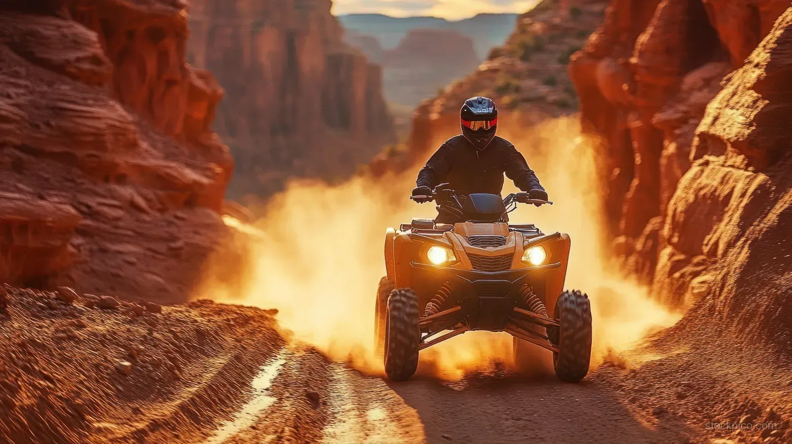 Helmeted rider speeds an orange ATV through a dusty narrow red-rock canyon at sunset with a dust cloud behind.