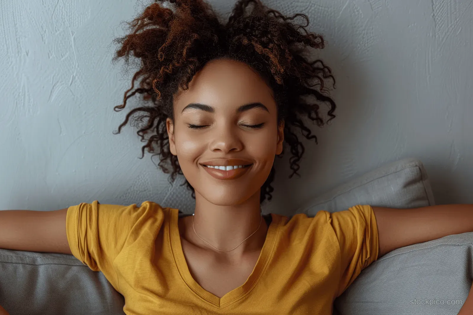 A smiling woman stretches on a couch in a bright living room with eyes closed.