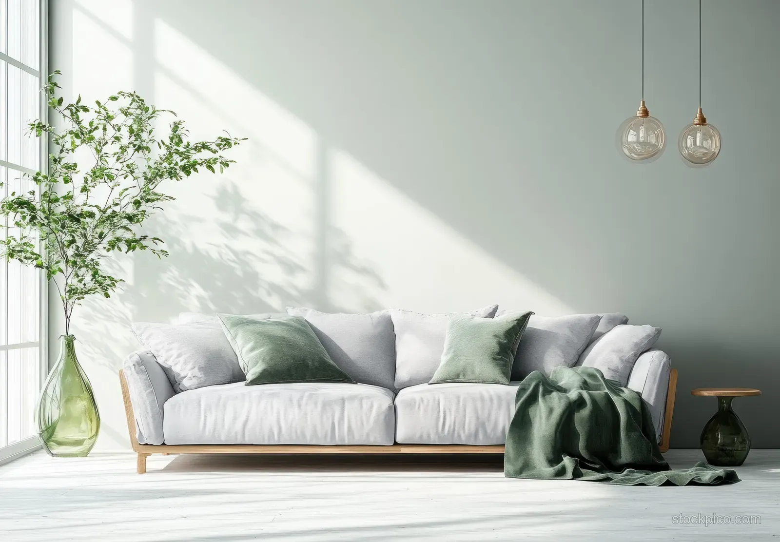 Light gray sofa with green throw and pillows beside a vase with leafy plant in sunlit minimalist room.