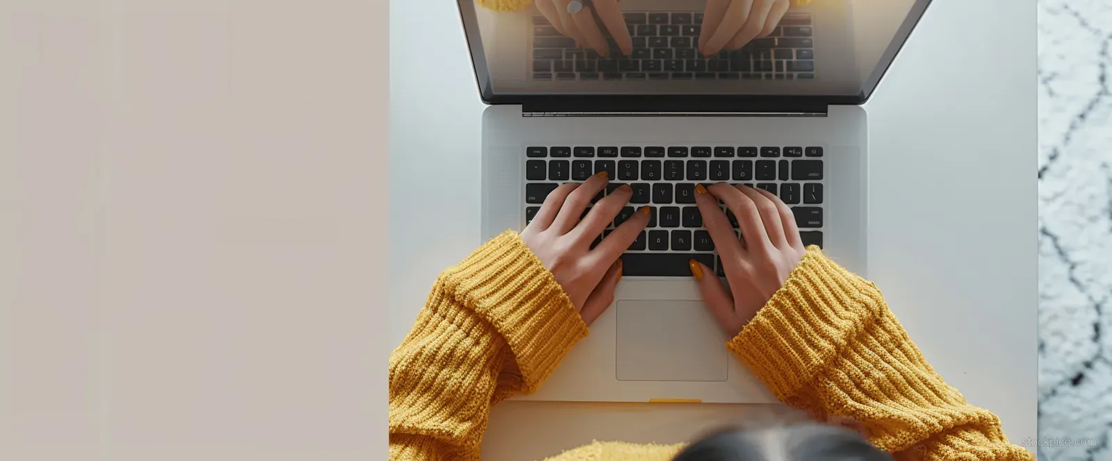 Woman’s hands typing on a laptop at a home office desk with warm sweater sleeves visible.