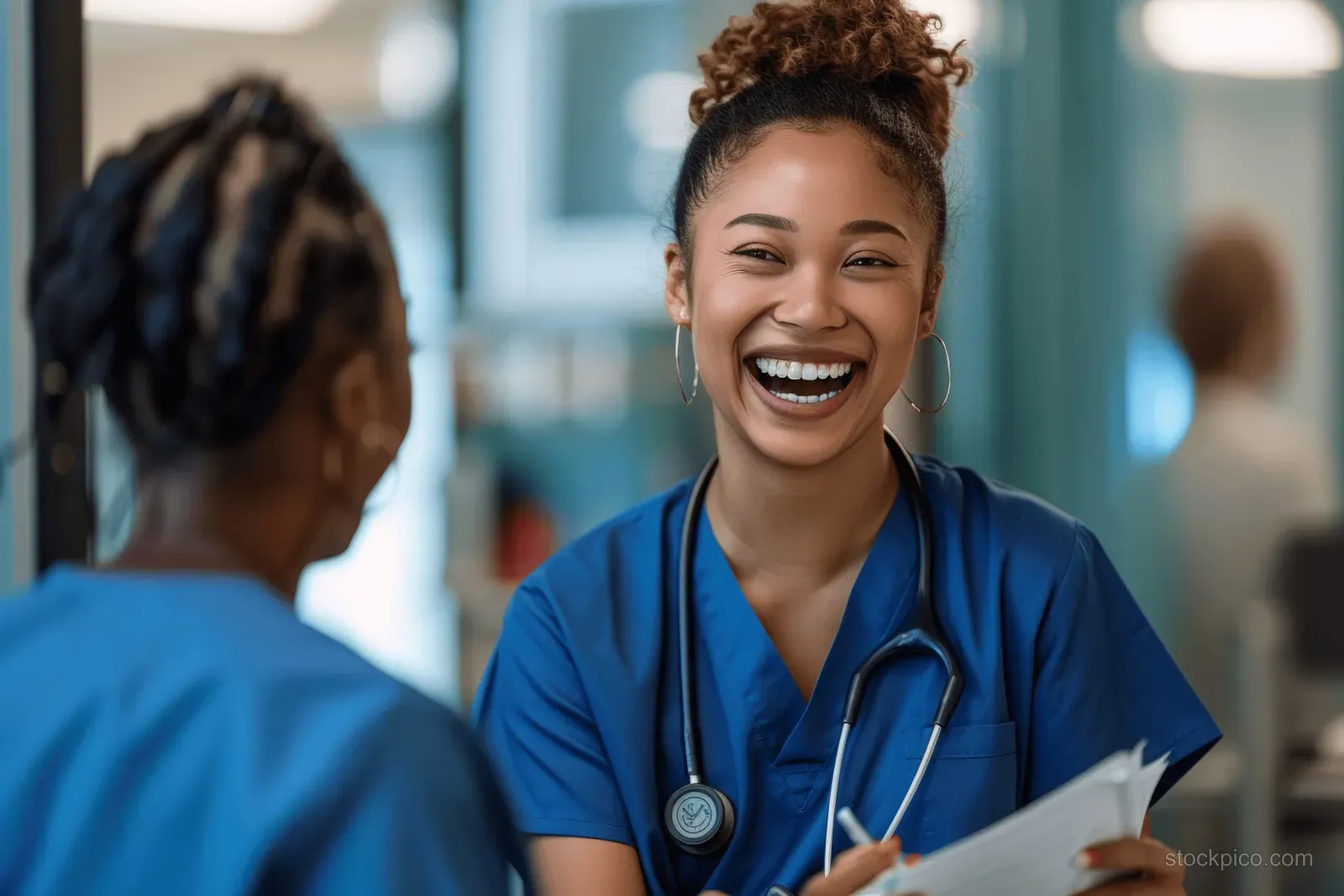 A smiling nurse in blue scrubs talks with a patient in a hospital hallway while holding a clipboard.