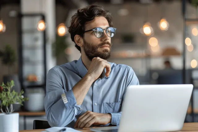 Focused businessman with glasses analyzing laptop at office desk, hand on chin