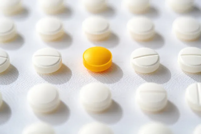 A bright yellow round pill stands out among multiple white round tablets on a white surface.