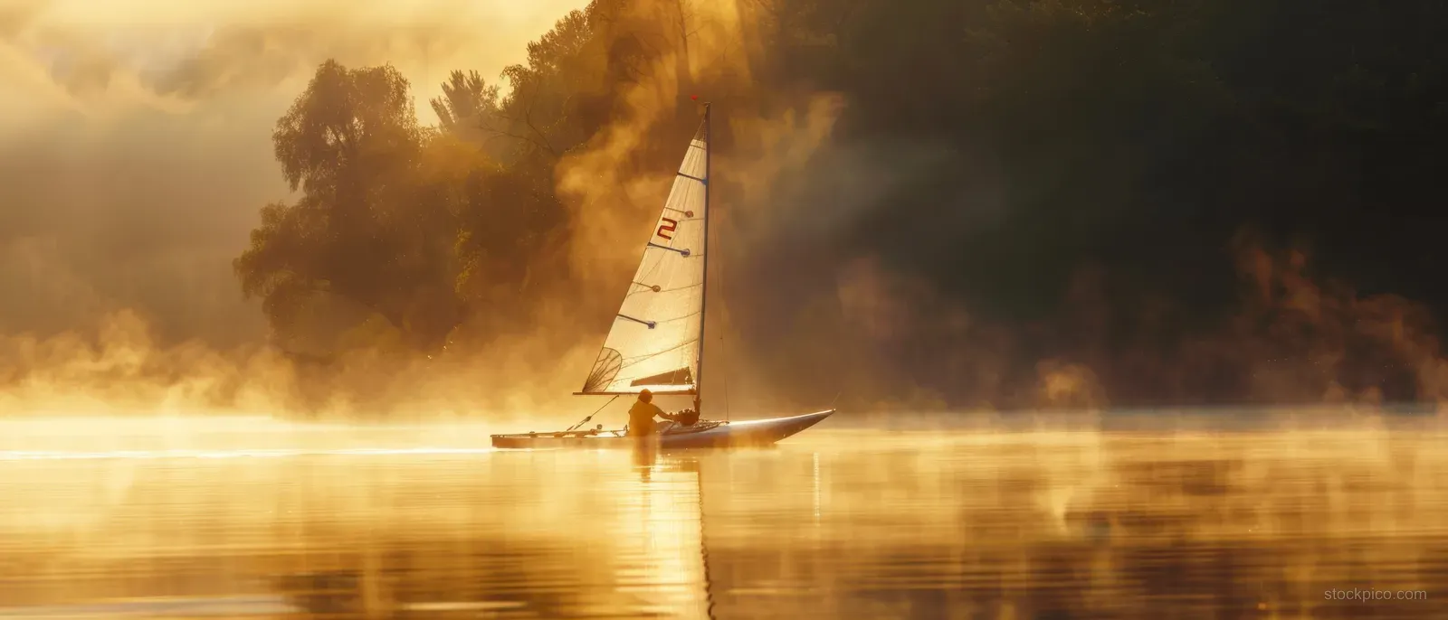 A Nacra 17 catamaran sails through sunrise mist on calm water, casting golden reflections behind the mast.