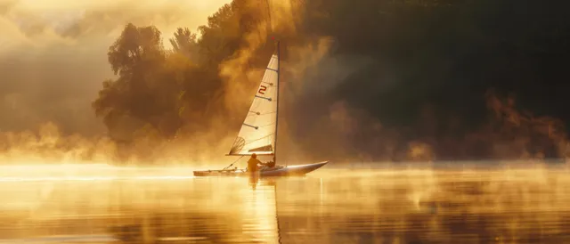 Nacra 17 catamaran sailing across misty Canadian water at sunrise with golden reflections