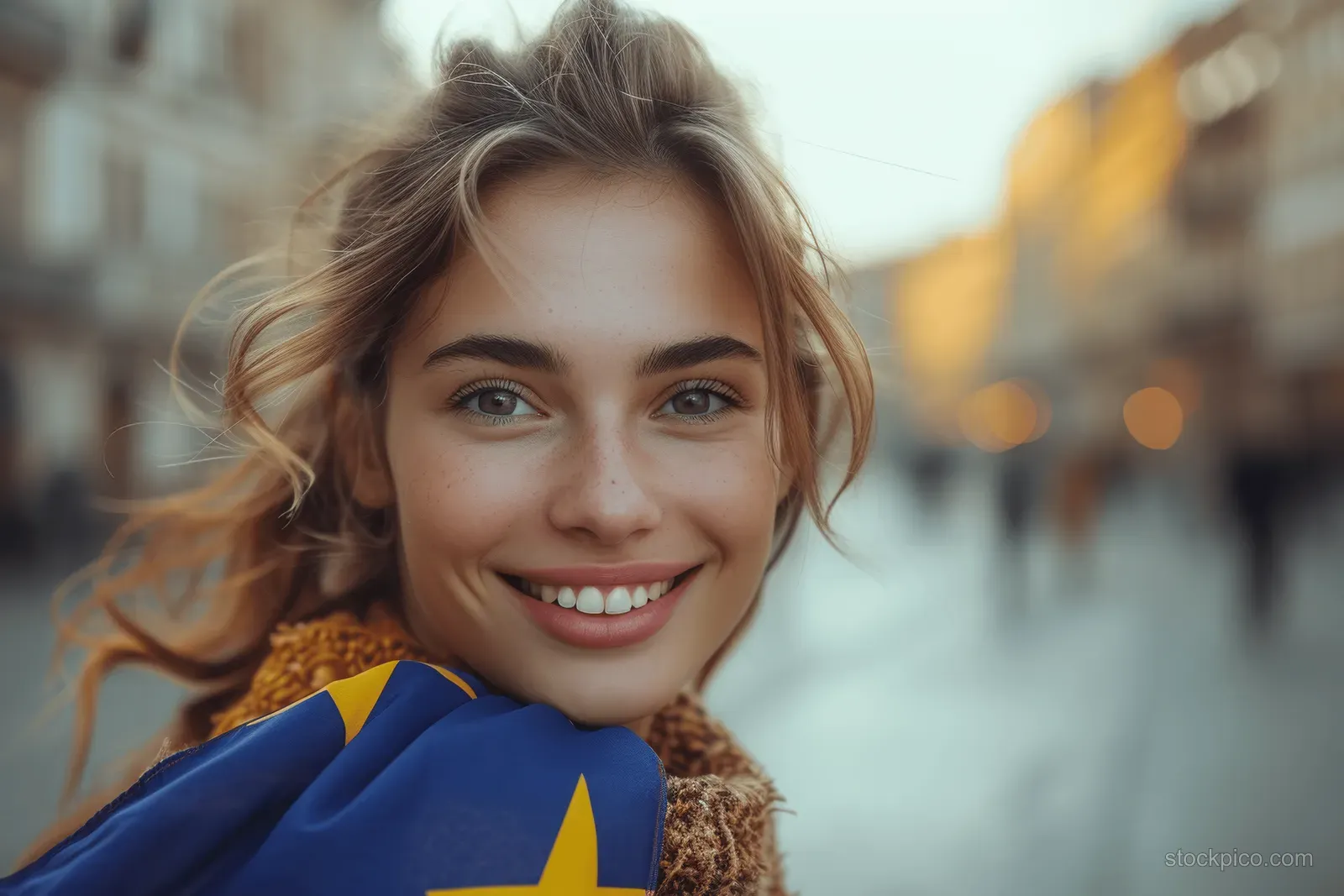 Young woman waves a European Union flag in a historic street with warm sunset light and blurred passersby.
