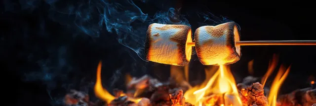 Two marshmallows on sticks roast over a campfire with bright flames and drifting smoke in the dark.