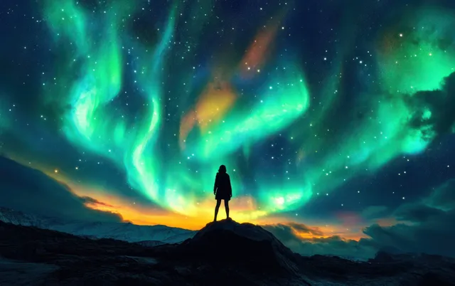 Silhouette stands on rocky ridge beneath vibrant green-blue aurora and starry mountains at dusk