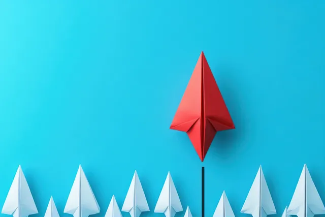 Red paper airplane rising above a row of white paper planes on blue background