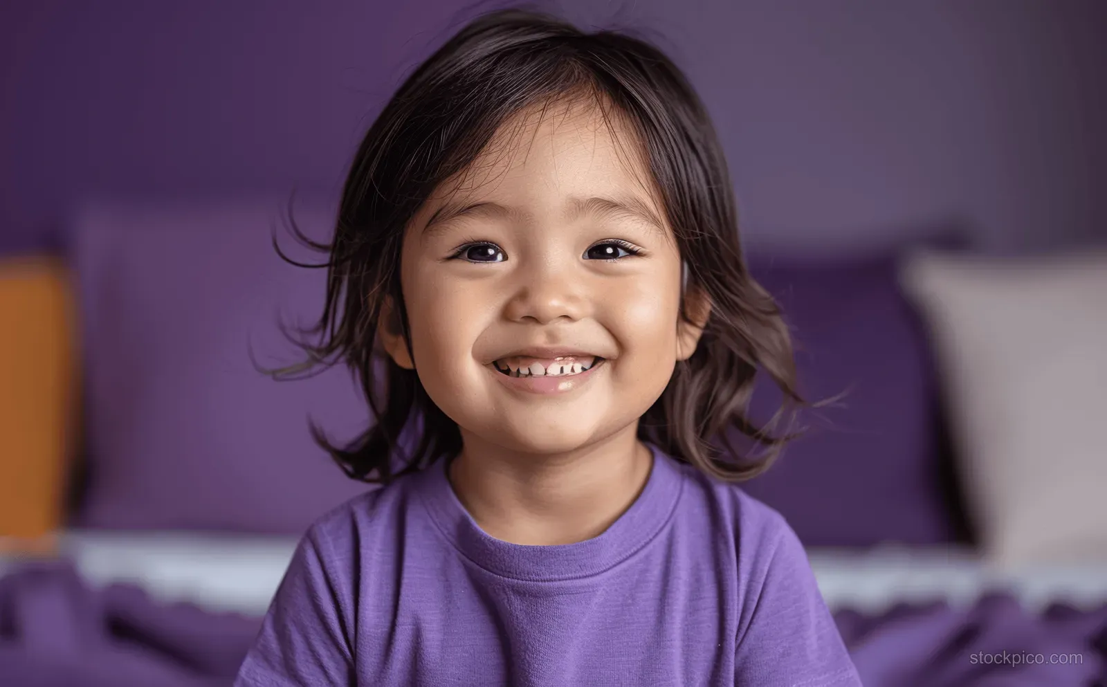 Asian toddler boy laughing in a purple T-shirt while seated on violet bedding in a close-up portrait.