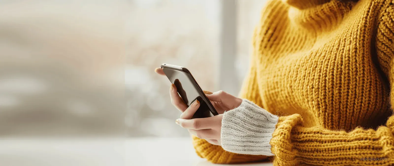 Young woman in a yellow sweater taps a smartphone screen at an office desk.