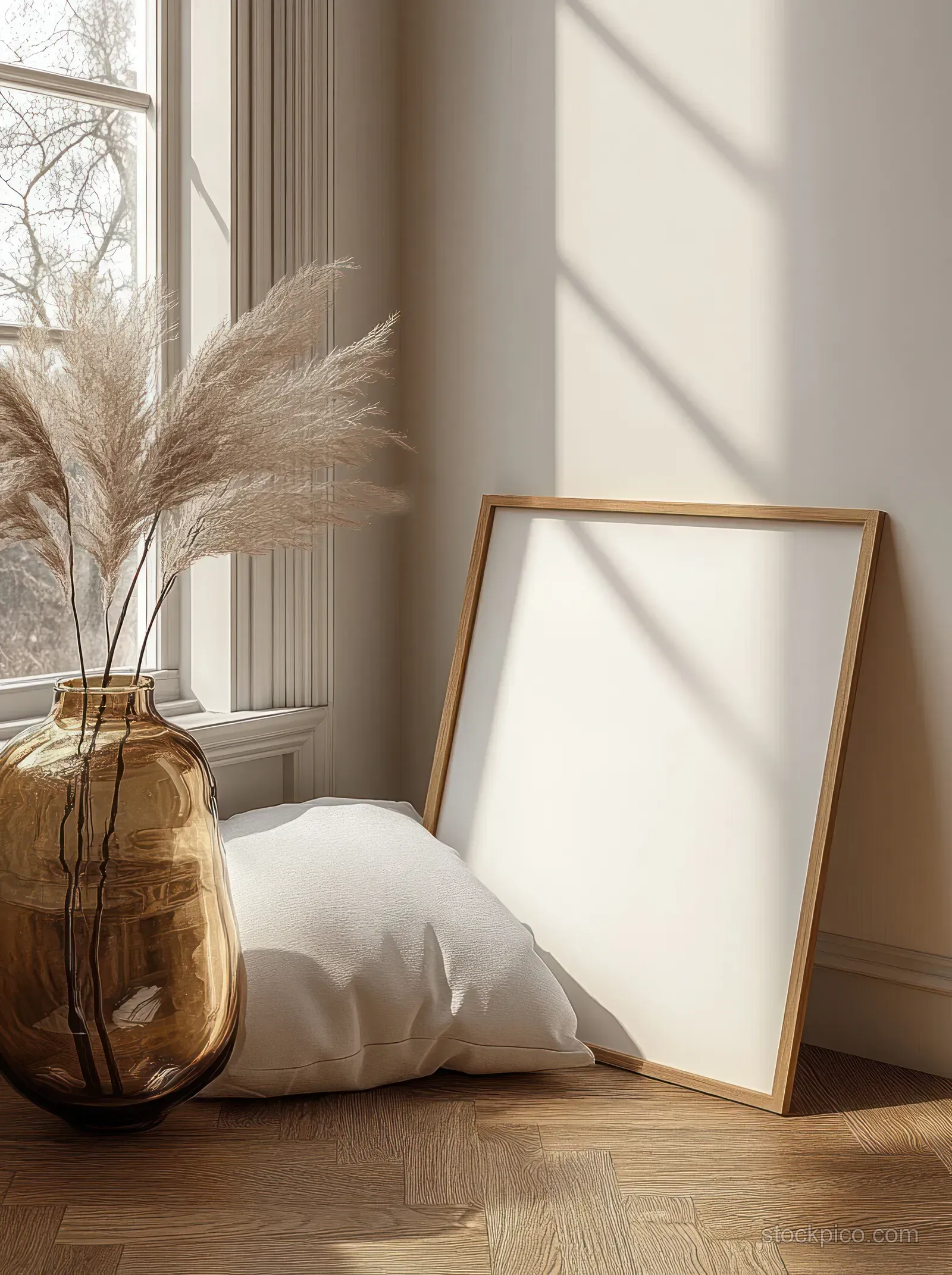 Blank picture frame leaning in a sunlit minimalist living room beside a white pillow and pampas grass vase.