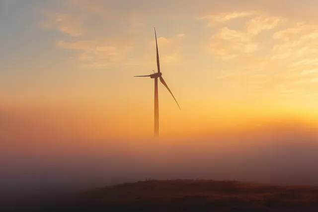 Lone wind turbine on a foggy grassy hill at sunset, blades cutting the misty air