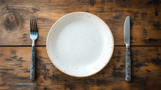 Empty speckled ceramic plate with fork and knife arranged on a rustic wooden table.