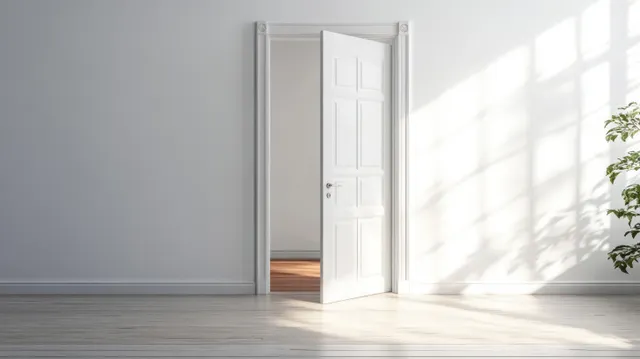 A half-open white door in a minimalist room lets bright sunlight glow onto the wooden floor.