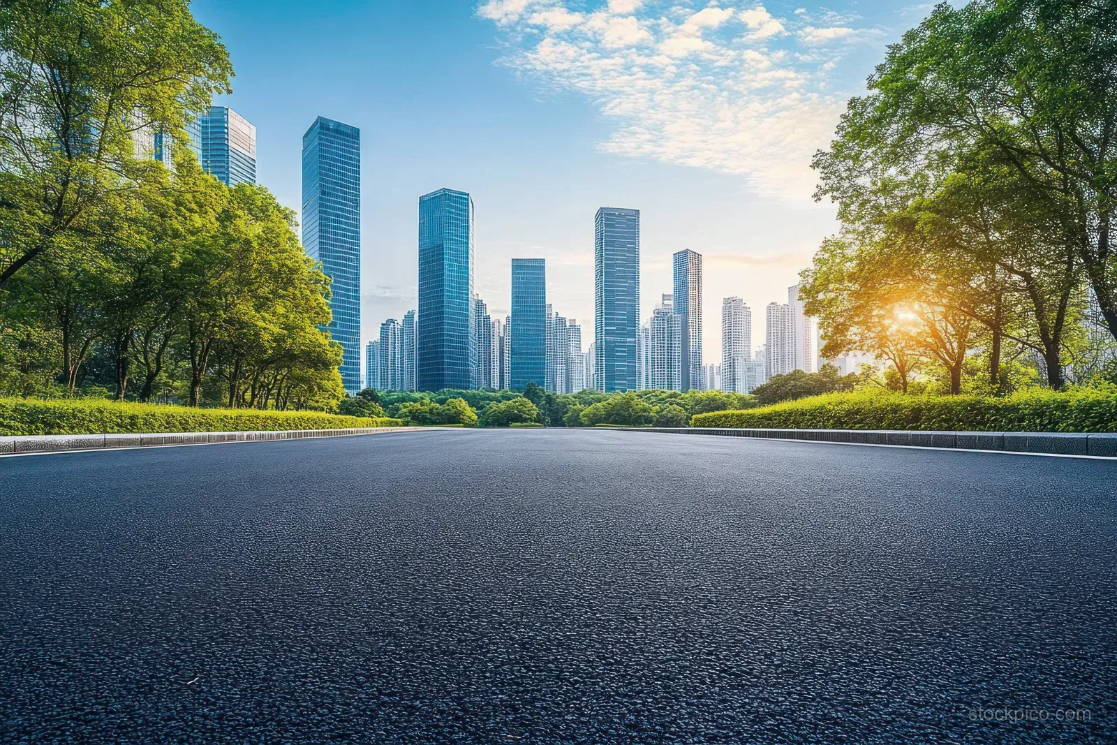 Empty wide asphalt road leads toward modern skyscrapers as sunrise glow lights trees along both sides.