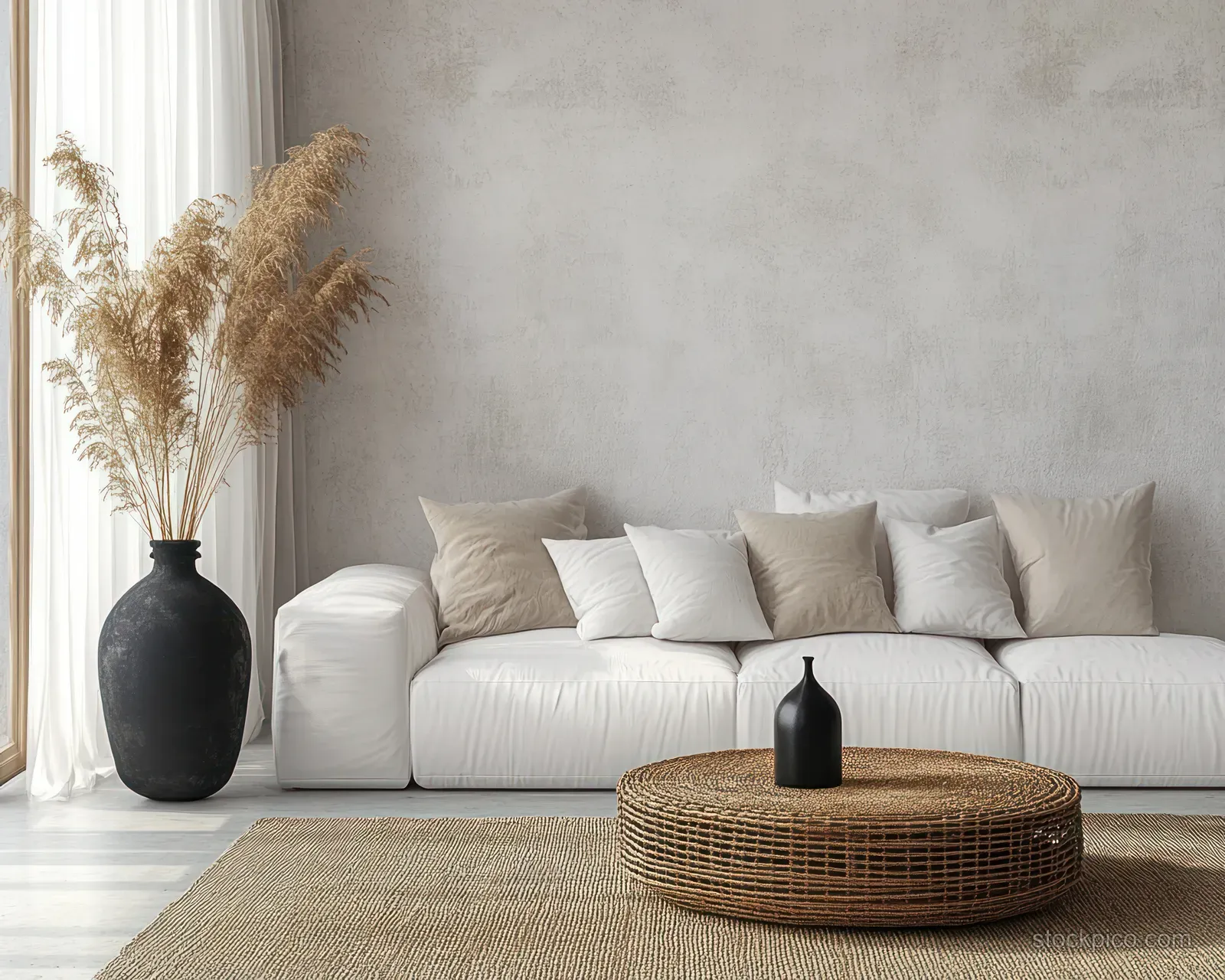 A white sofa and round rattan coffee table sit in a sunlit minimalist living room with a black vase and dried grasses.