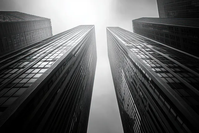 Low-angle black and white view of towering skyscrapers with reflective glass facades converging into a bright overcast sky.
