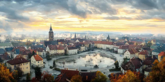 Panoramic autumn view of a historic town with a winding river, red rooftops, and church towers in golden light.