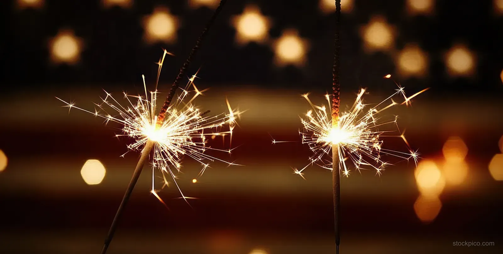 Two sparklers with American colors glow against a dark background with warm blurred lights.
