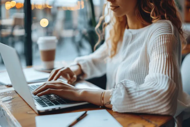Woman in white shirt types on a laptop at a cafe table with documents and a pen visible on the desk.