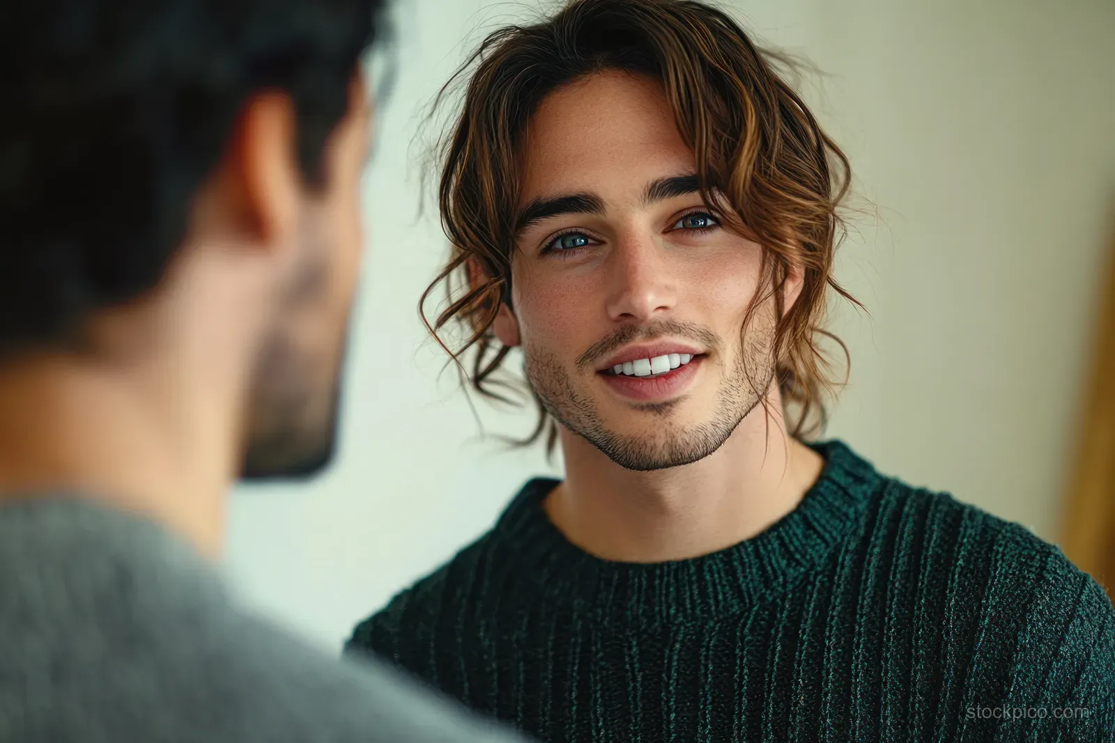 A smiling man in a dark knit sweater talks with a coworker in a softly lit office meeting room.