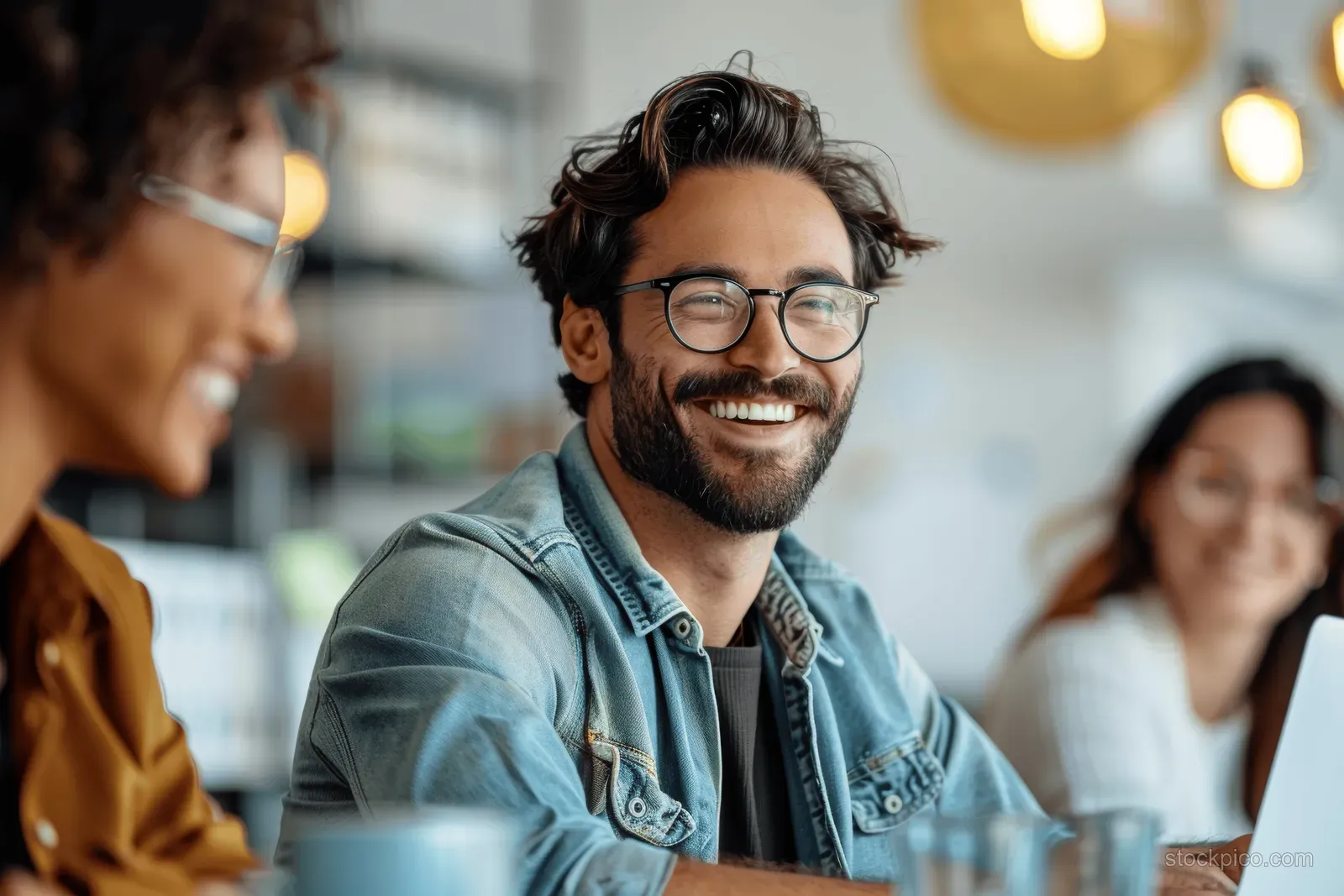 Smiling man with eyeglasses typing on a laptop at an office table during a meeting with coworkers behind him.