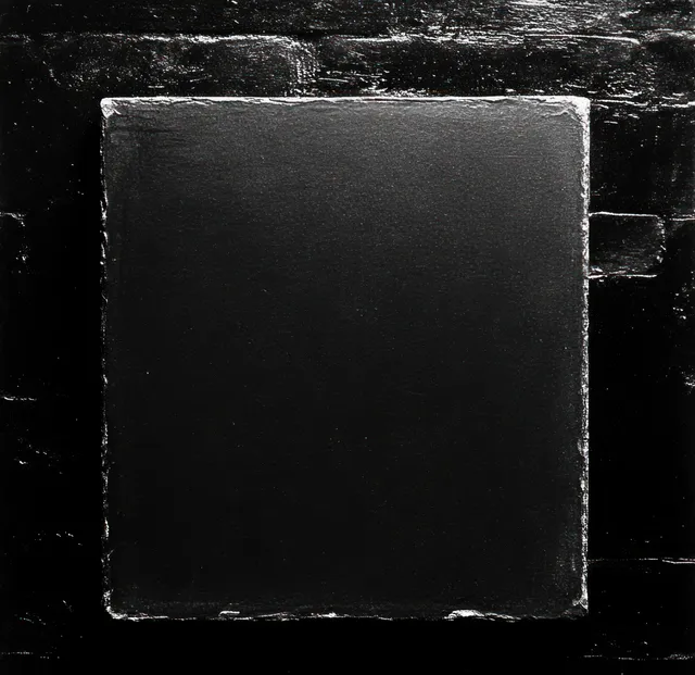 A rough black square metal plate is mounted on a dark brick wall, with worn scratched edges.