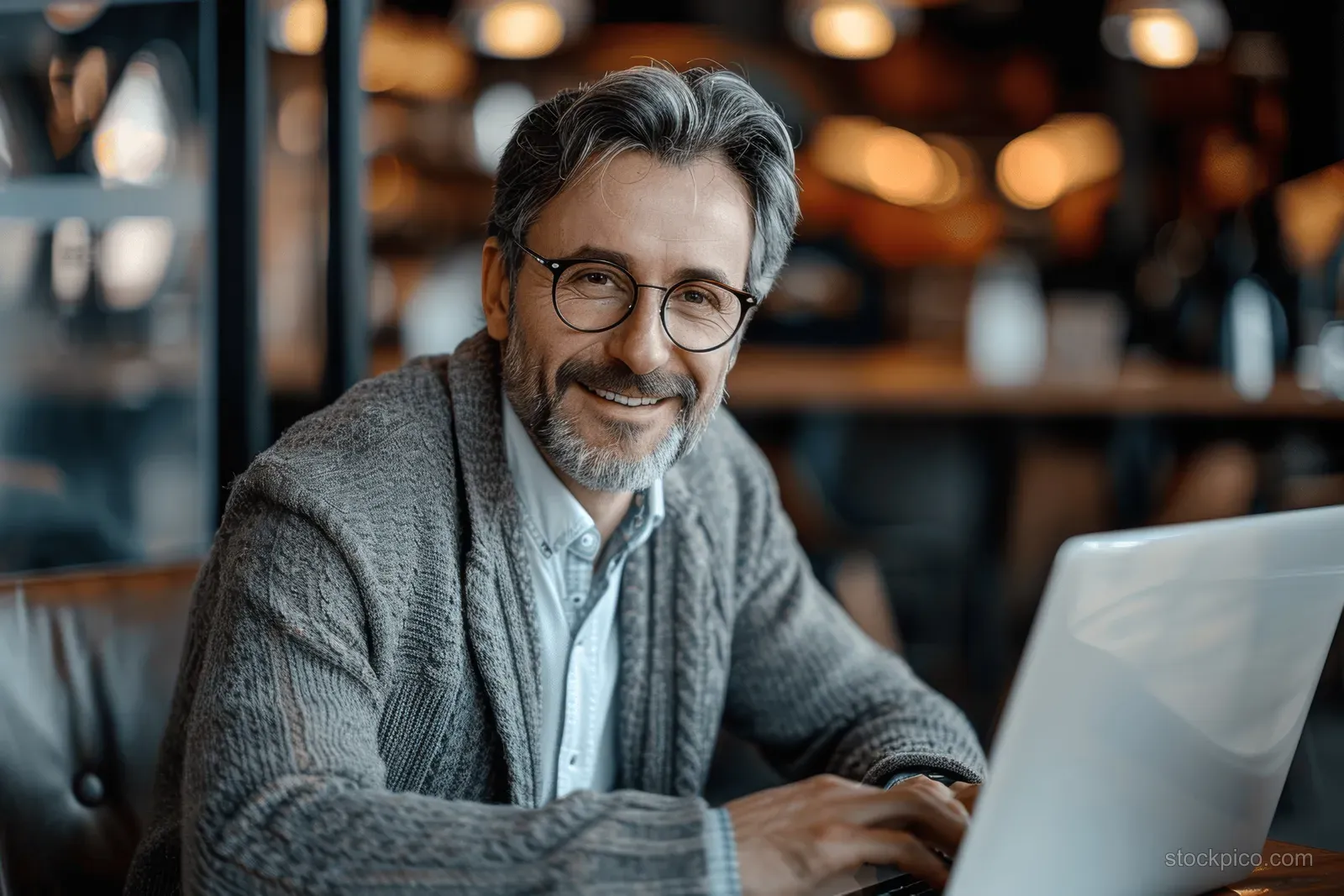 Smiling businessman wearing glasses, sweater, and tie typing on a laptop at an office café counter.