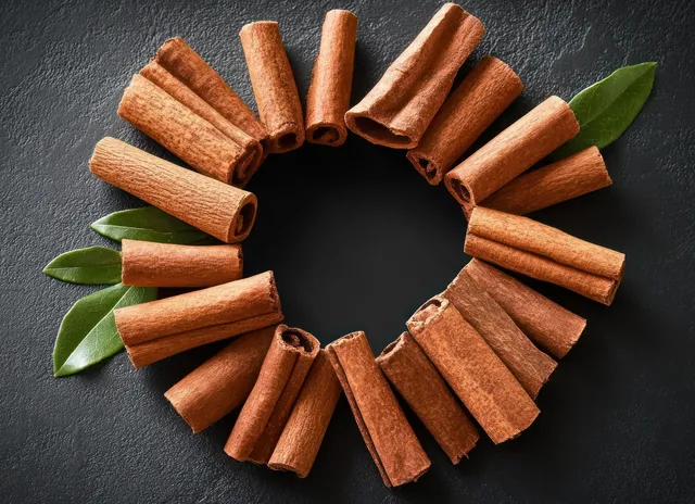 Cinnamon sticks arranged in a circular ring with green leaves on a dark textured background.