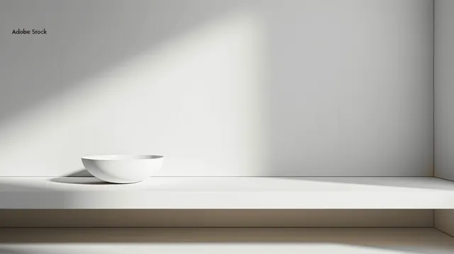 Minimalist white bowl sits on a white shelf against a light gray wall with soft sunlight shadows.