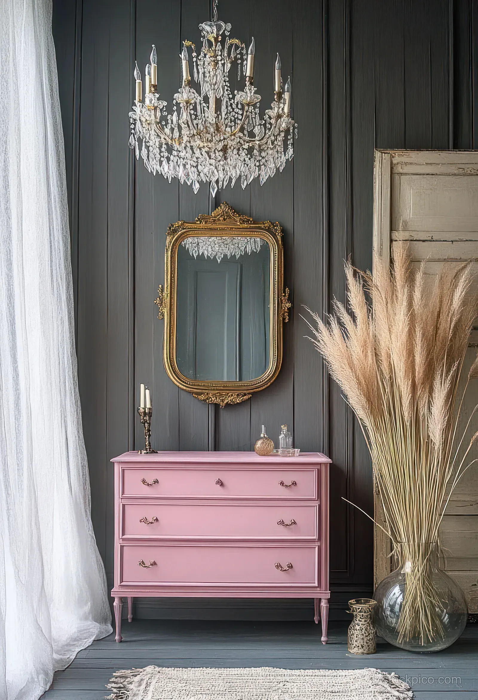 Pink chest of drawers under a crystal chandelier with a gold mirror on dark paneling and pampas grass in a glass vase.