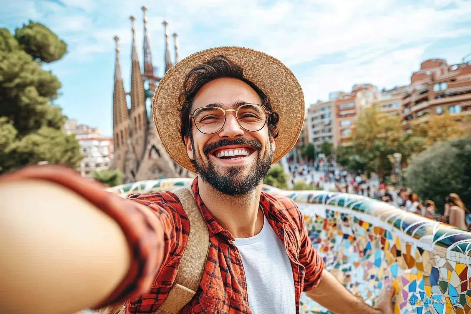 Smiling man taking a selfie at Park Güell with colorful mosaic terrace and distant Sagrada Família.