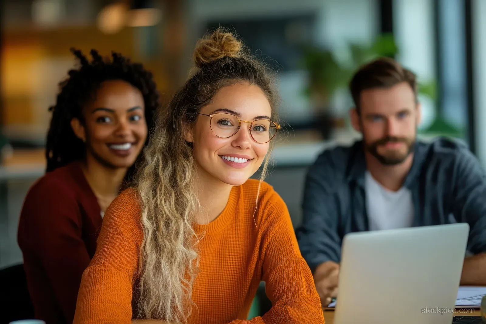 A young woman smiles at a laptop while coworkers sit behind her in a bright office meeting.