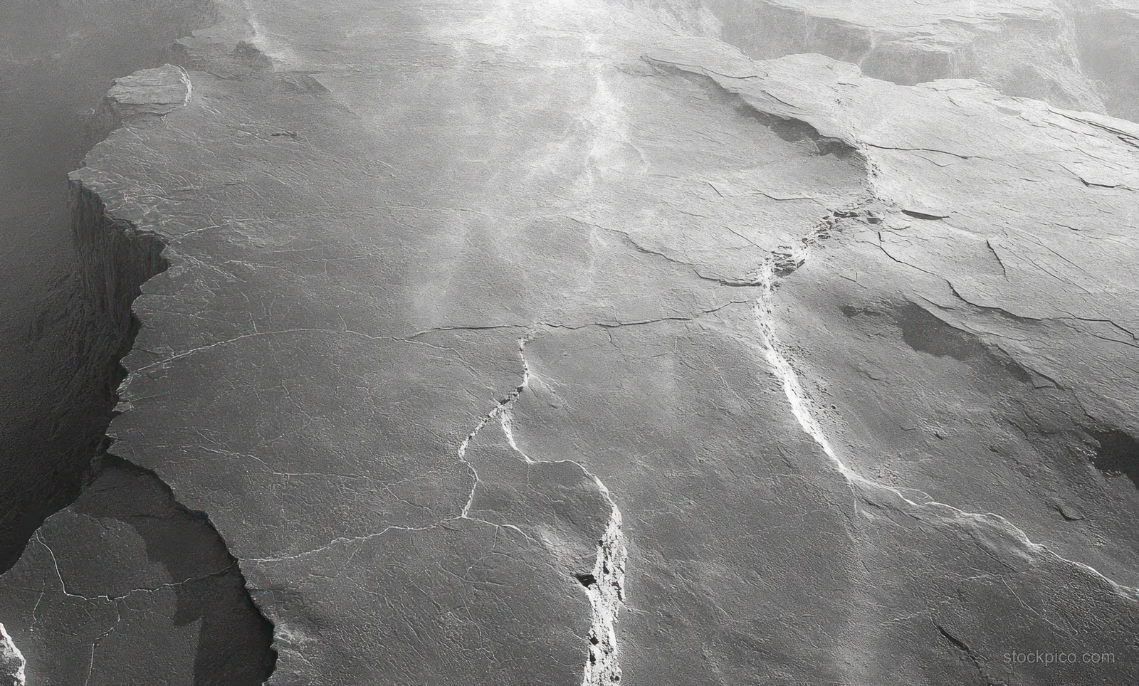 Weathered grey rock outcrop with deep cracks and fissures spreading across an uneven surface under soft diffused light.