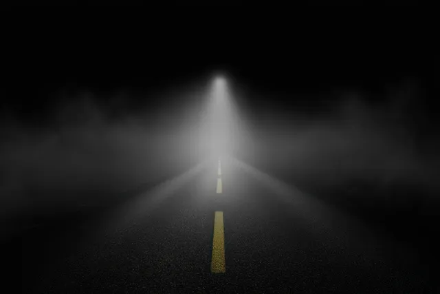A deserted road at night fades into thick fog, lit by one distant car headlight and yellow center lines.
