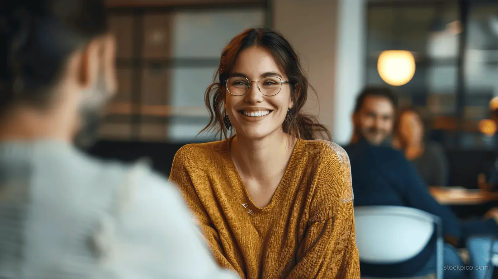 Woman with glasses and a yellow sweater smiles during an indoor conversation while people sit behind her.