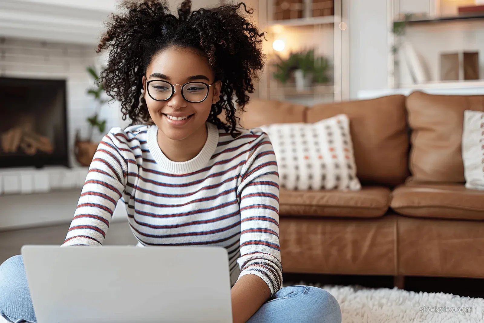 Smiling woman in striped sweater and glasses using a laptop on a couch in a home living room.