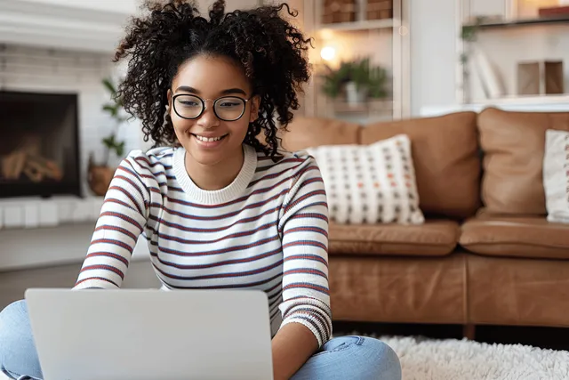 Smiling woman using a laptop in a living room, striped sweater and glasses