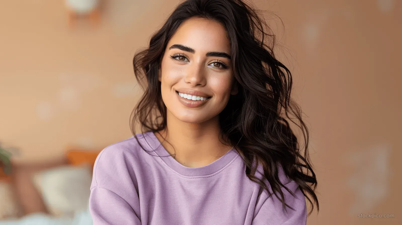 A young woman with long dark hair smiles while wearing a purple sweatshirt in soft natural light indoors.