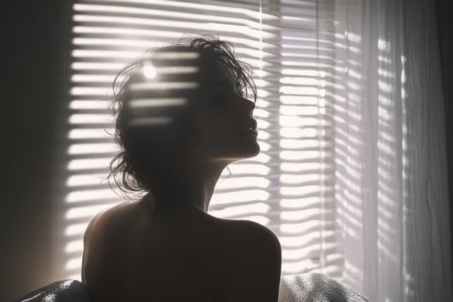 Silhouette of a curly-haired person sitting beside window blinds casting striped shadows in a dim room.
