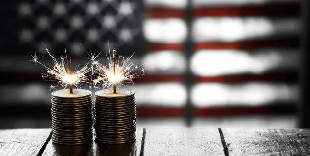 Two sparklers burn atop stacked coins with a blurred American flag in the background.