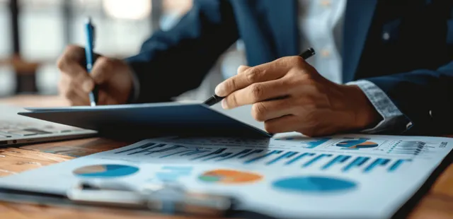 A businessman holds a pen over printed financial charts and documents on an office desk.