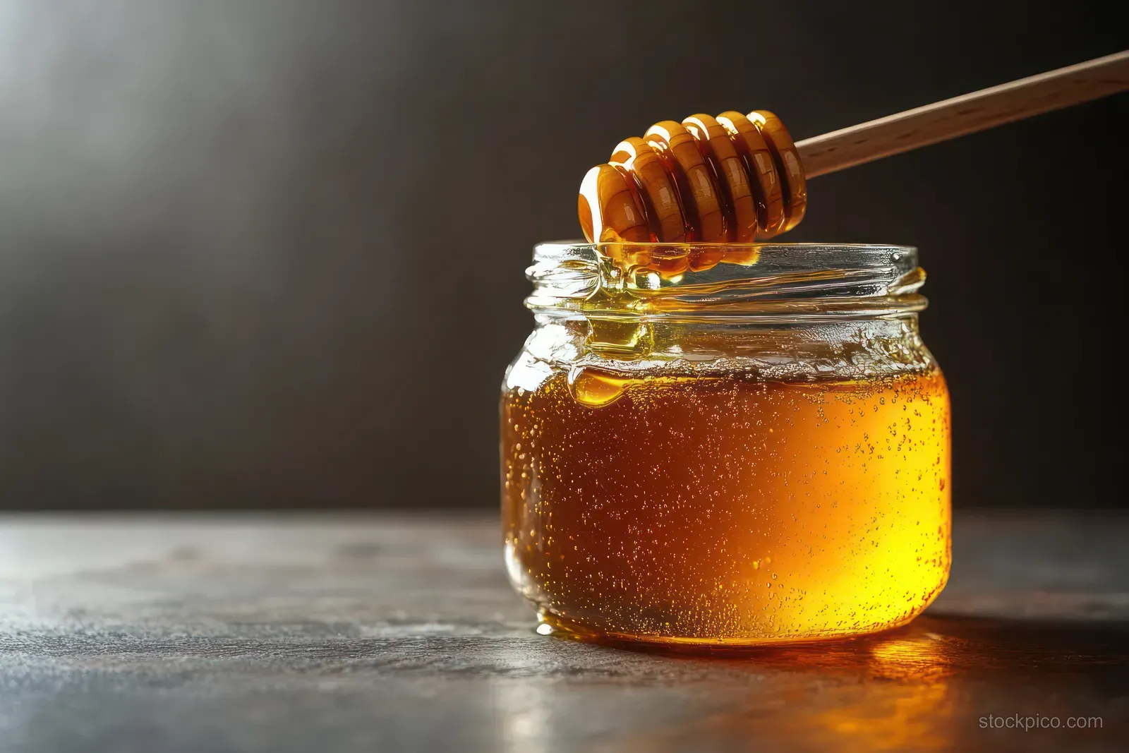 A glass jar of golden honey sits on a dark table, with a wooden dipper dripping honey onto the surface.